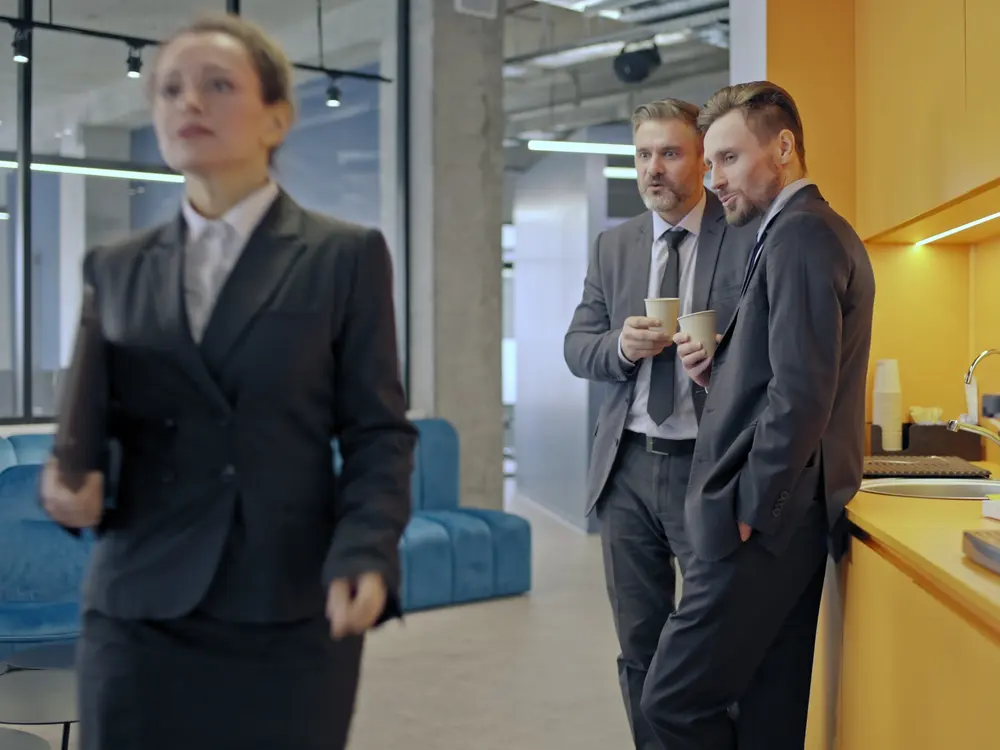 A woman walks past two male coworkers who watch her and whisper in an office break area.